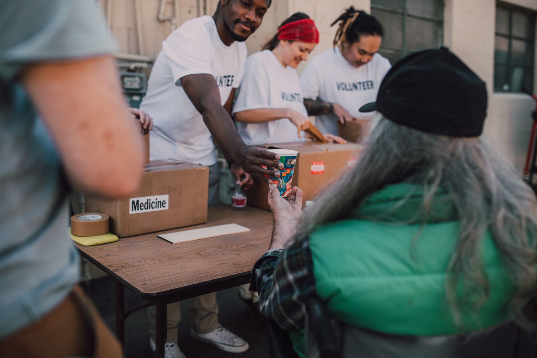Volunteers in white T-shirts hand out supplies from a table stacked with cardboard boxes