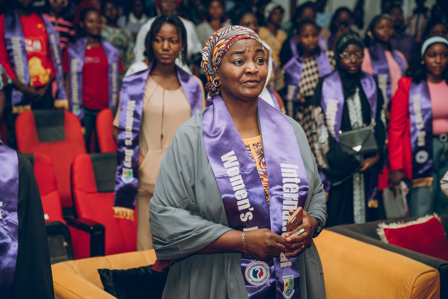 A woman wearing an International Women's Day sash stands among other women in sashes at an indoor ceremony