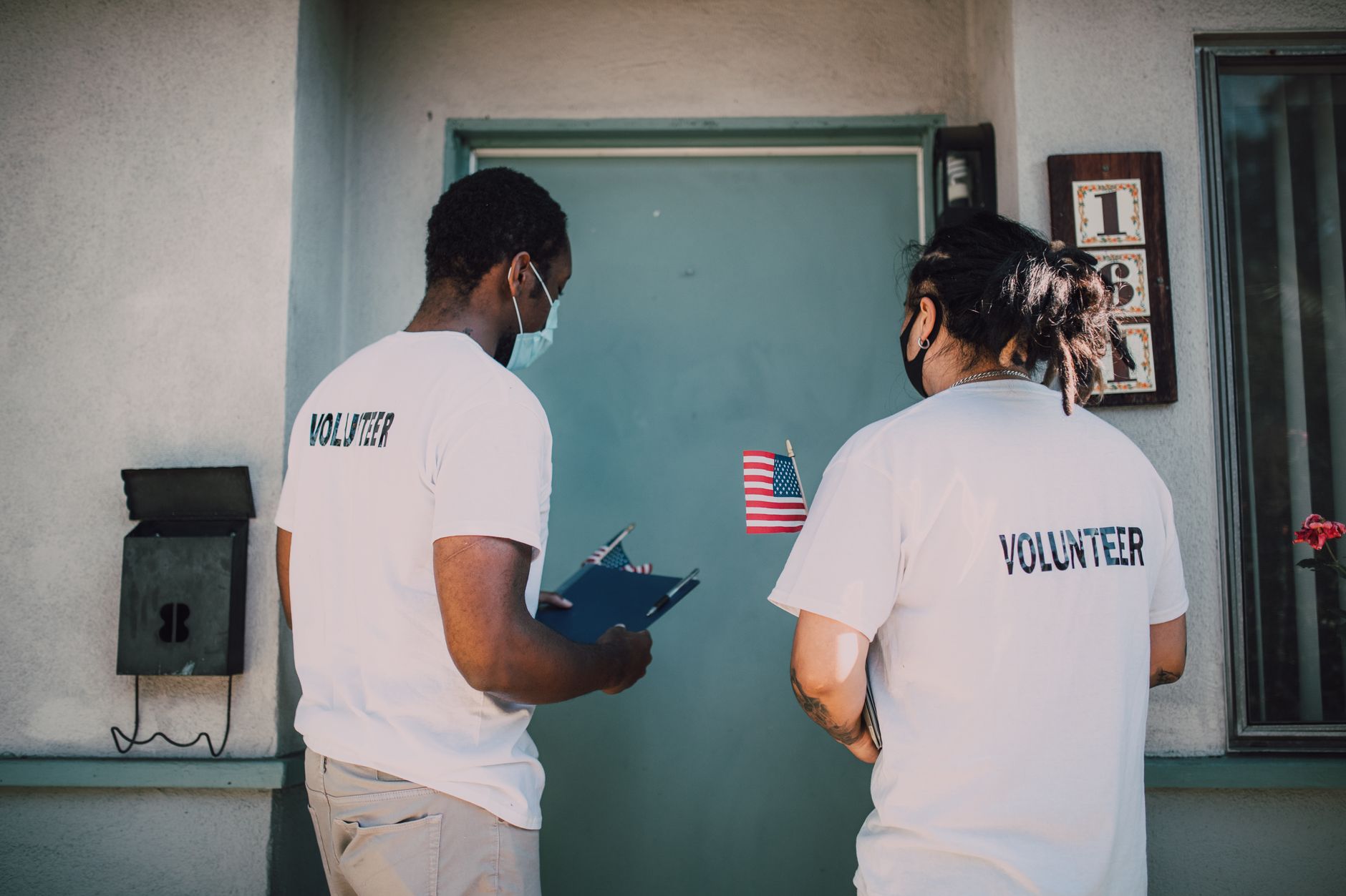 Two volunteers in face masks and volunteer T-shirts knock at the door of a house, holding clipboards