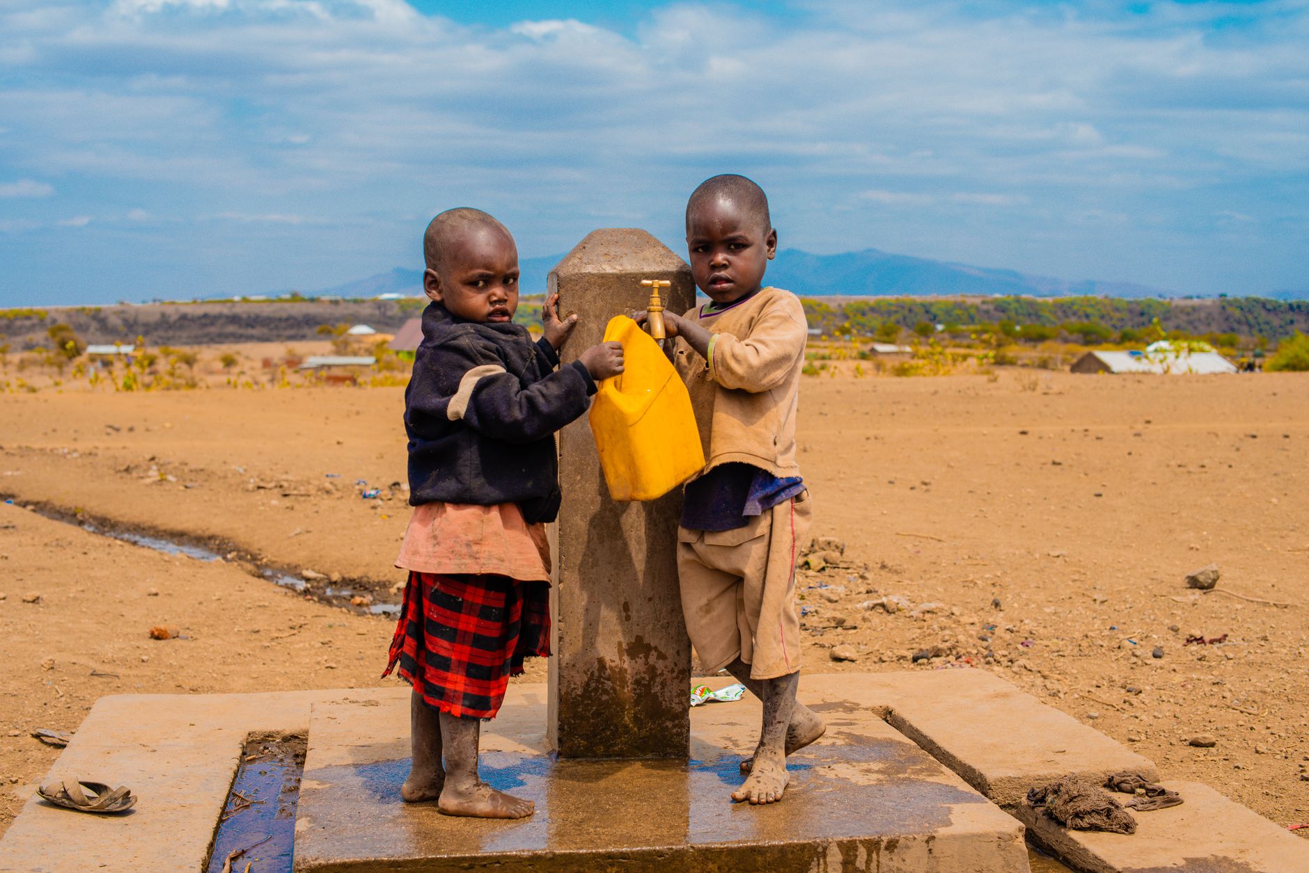 Two small children fill a yellow jerrycan at a standpipe in a dry landscape