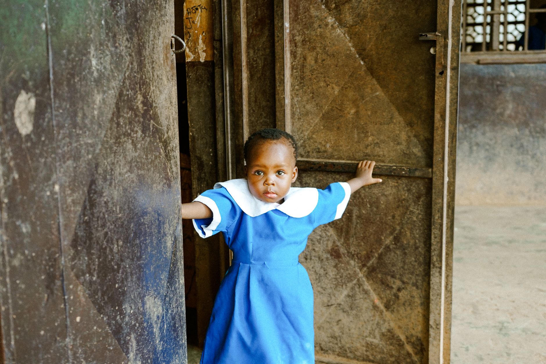 A young girl in a blue school uniform stands in a metal doorway