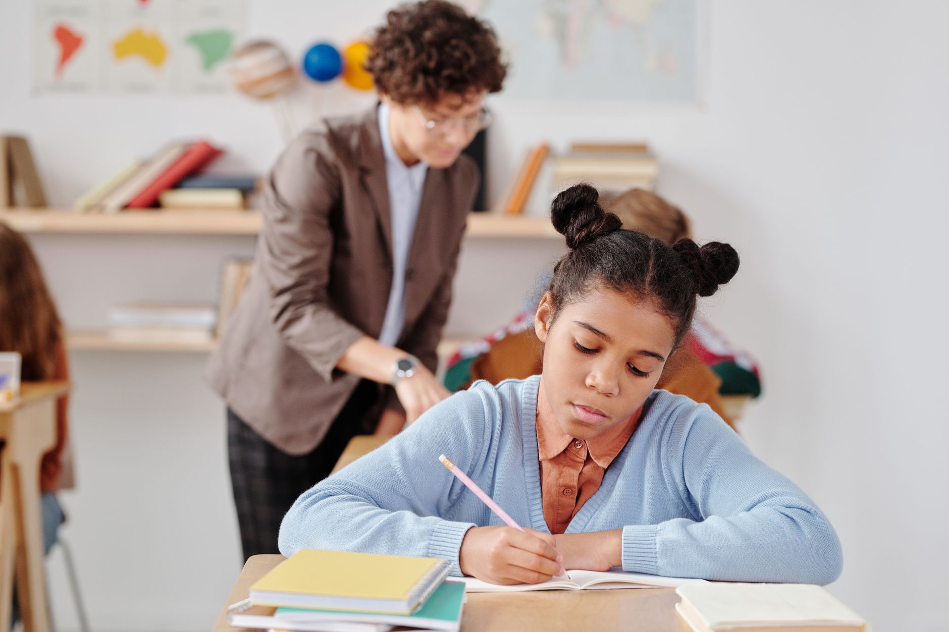 A girl writes in her exercise book at a classroom desk while a teacher looks on