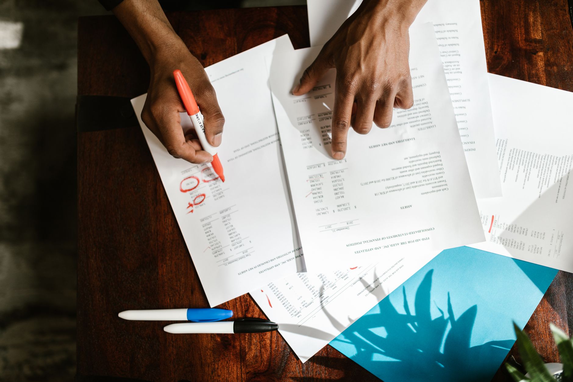 Two hands mark up printed documents with an orange pen on a wooden desk