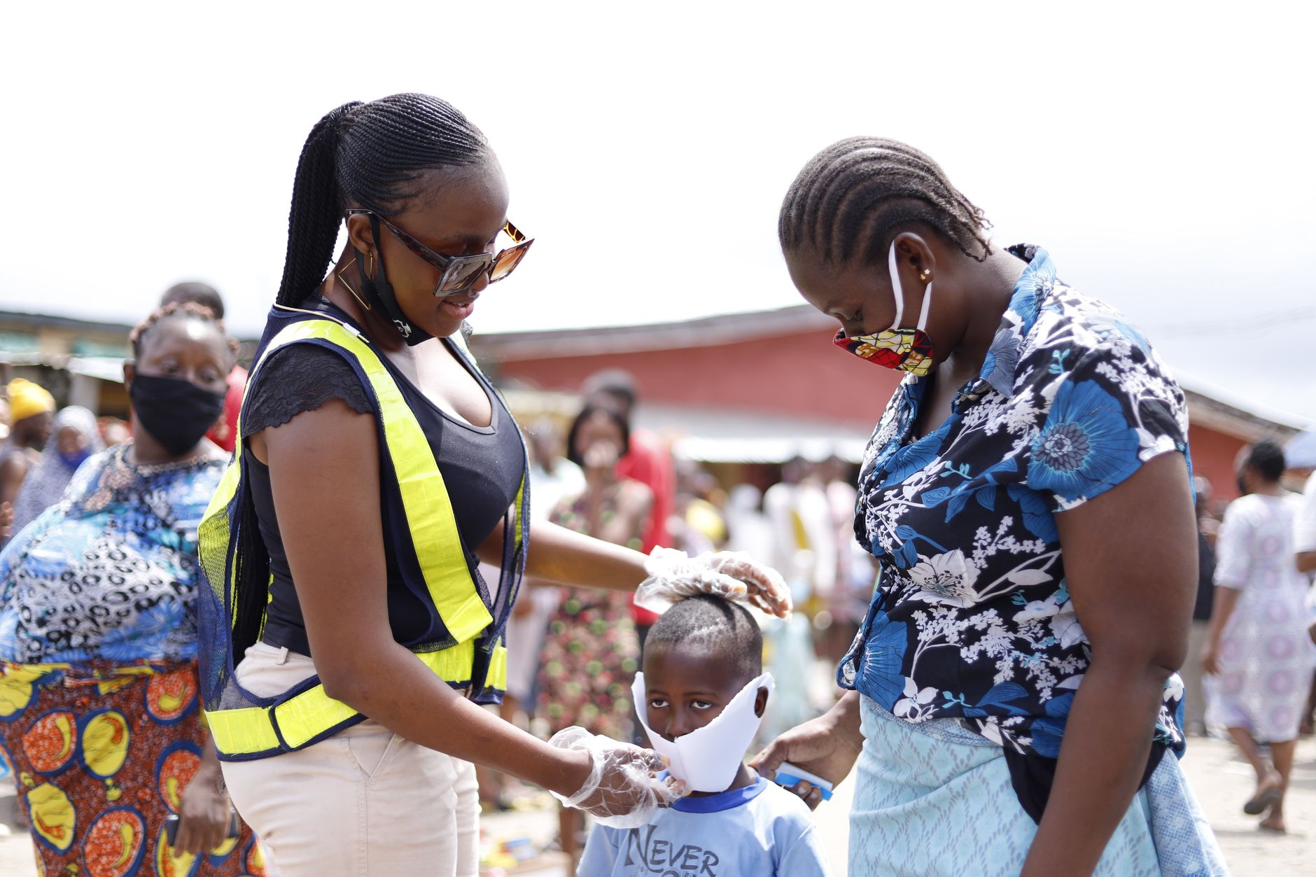 A health worker in a high-visibility vest fits a face mask on a child at a busy market
