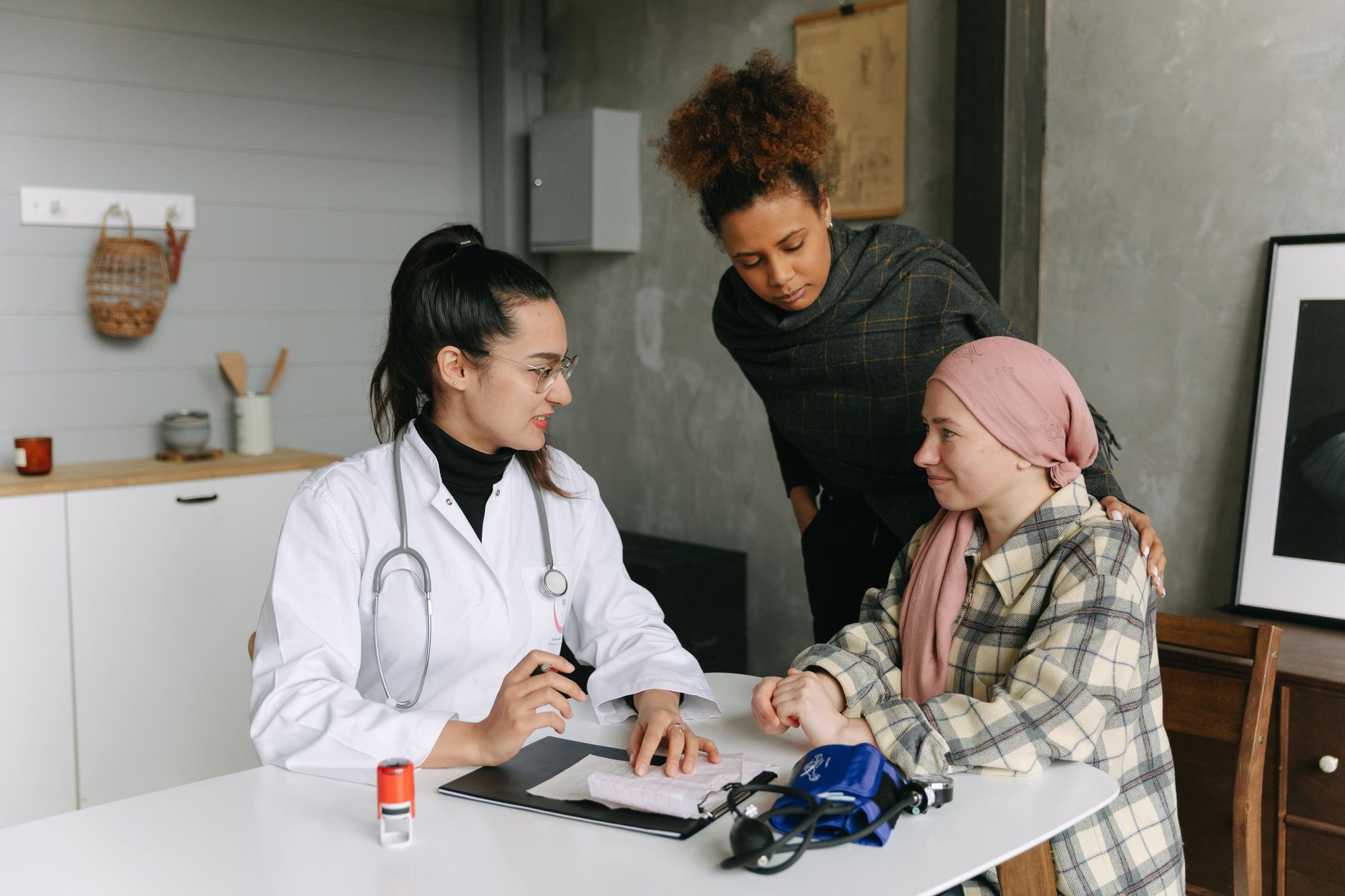 A doctor speaks with a woman in a headscarf at a clinic table while a companion rests a hand on her shoulder