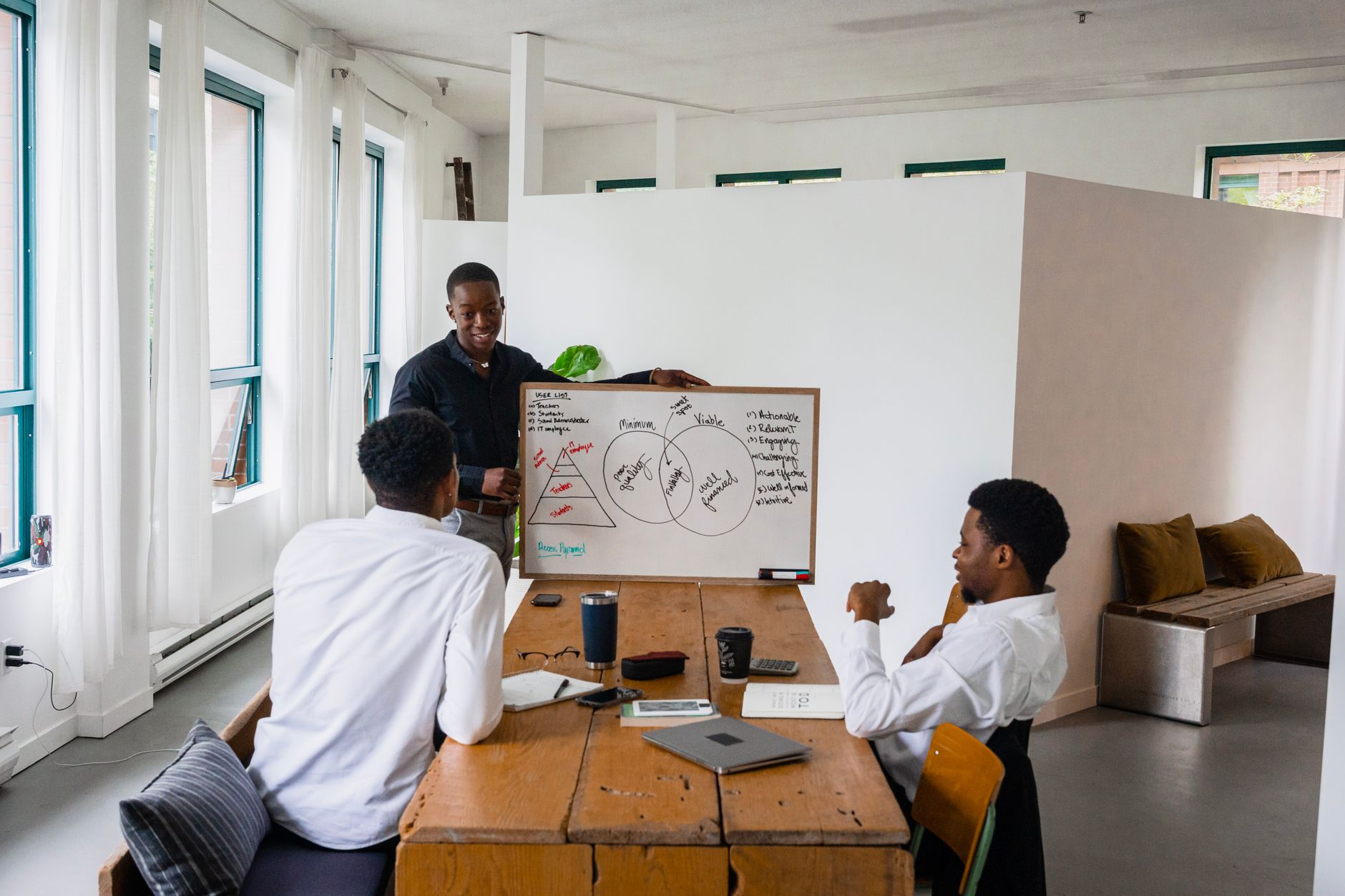 A young man presents a diagram on a whiteboard to two colleagues at a wooden table