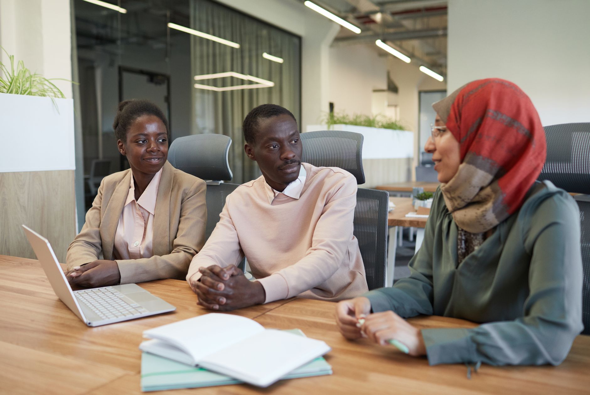 Three colleagues talk around an office table with a laptop and notebooks