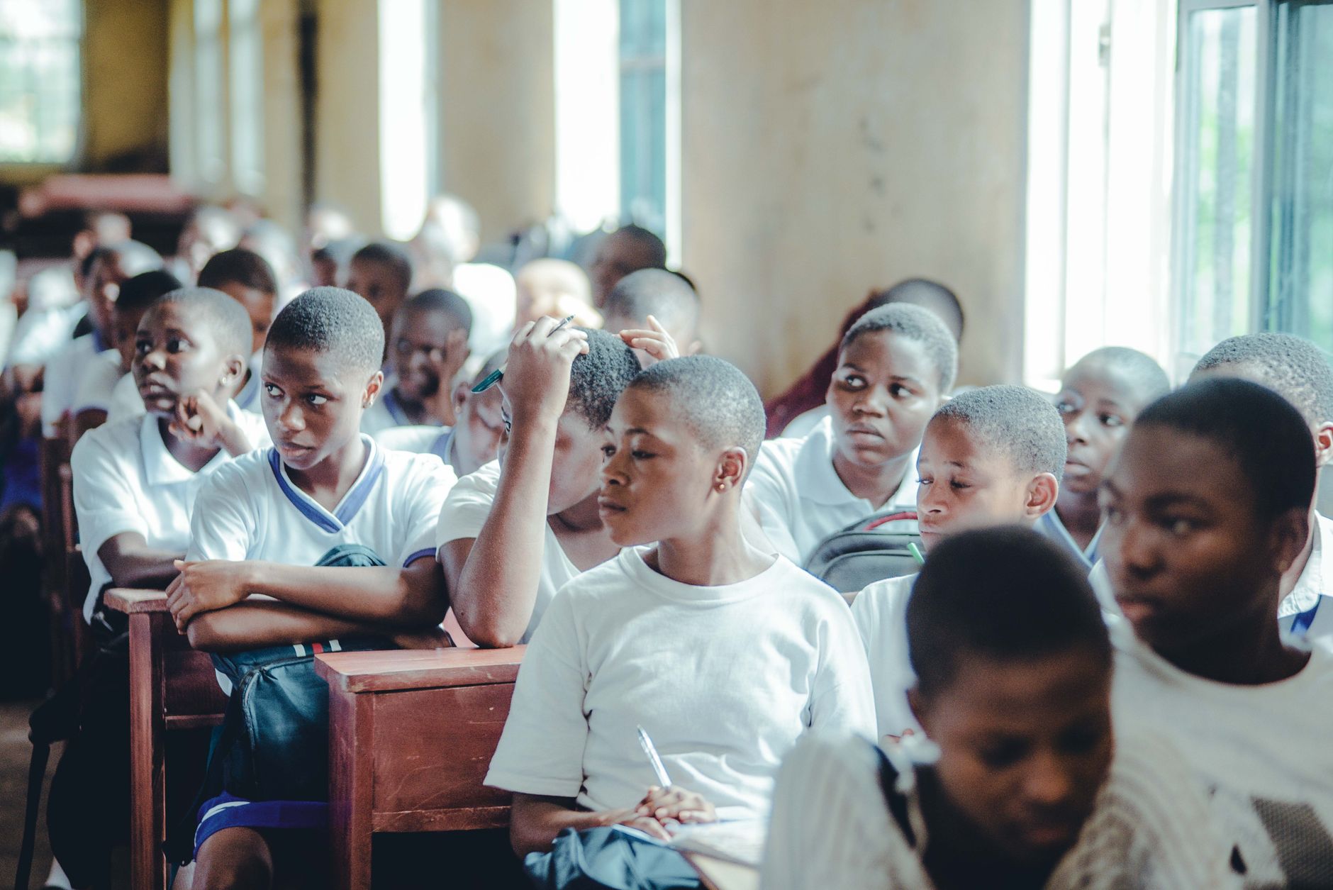 A crowded classroom of students in white school uniforms seated at wooden desks