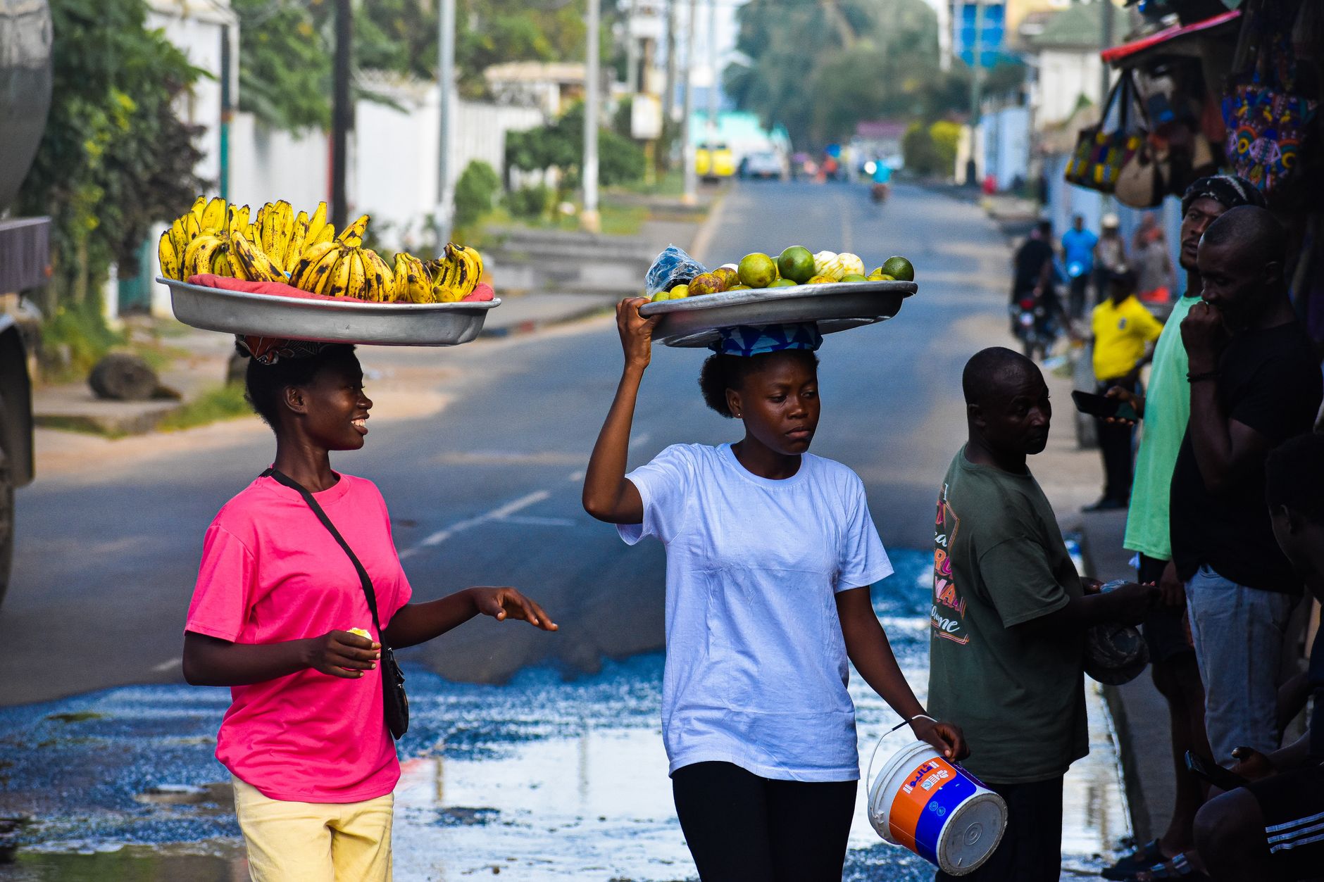 Two young women carry trays of bananas and fruit on their heads along a busy street