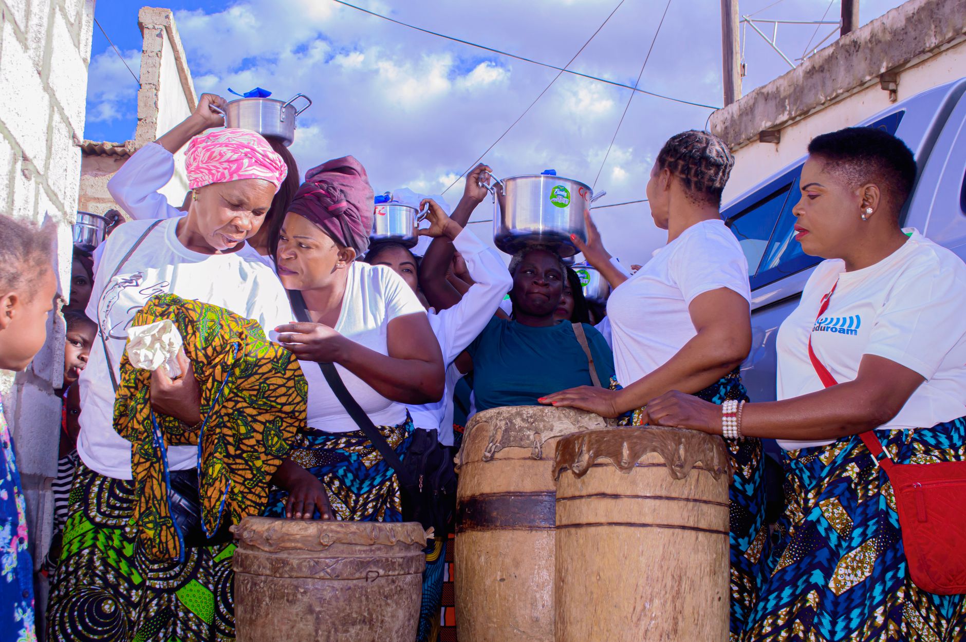 Women in white T-shirts and patterned wrappers carry cooking pots on their heads beside traditional drums