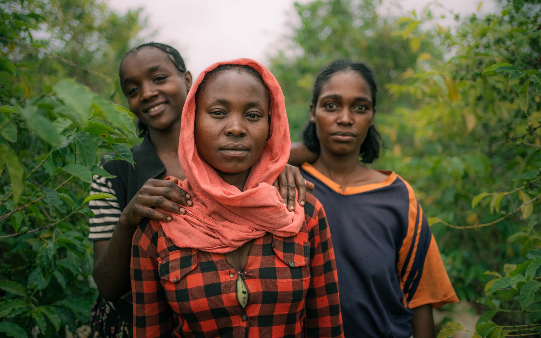 Three young women stand together among green plants, two resting a hand on the shoulders of the third