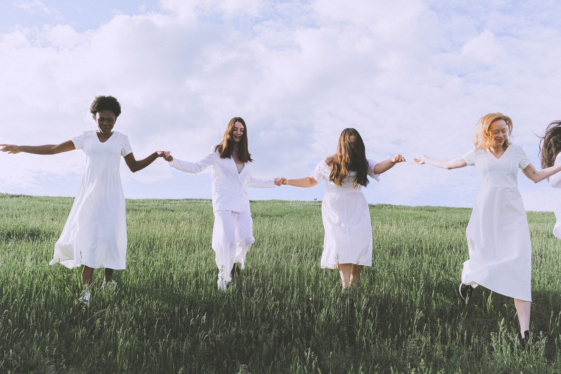 Four women in white dresses hold hands as they walk across a grassy field